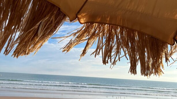 Een strandparasol met rieten dak, een zandstrand en de oceaan op de achtergrond.
