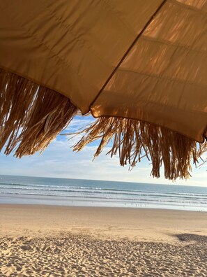 A beach umbrella with a thatched top, a sandy beach, and the ocean in the background.