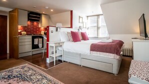 A bedroom with a bed, bedside table, and a small desk. There is a kitchen area with a stove and cabinets.