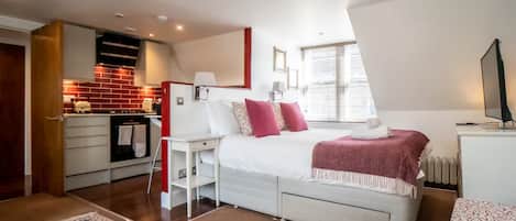 Un dormitorio con cama, mesita de noche y un pequeño escritorio. Hay una zona de cocina con estufa y armarios.