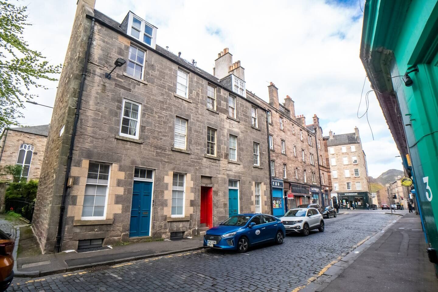 A street with historic stone buildings, a cobblestone road, and parked cars.
