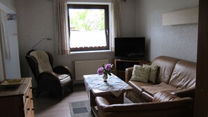 En stue med en sofa, en lænestol, et lille bord med en vase, et fjernsyn og et vindue med gardiner.