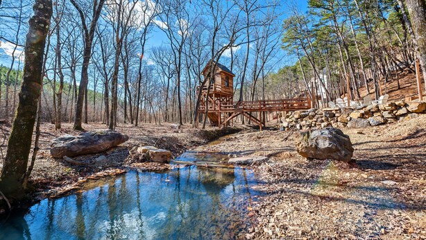 A rustic wooden cabin with a wooden deck, surrounded by a forest and a small stream.