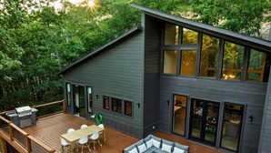 A modern house with a deck, a dining area, and a large window overlooking a forested area.
