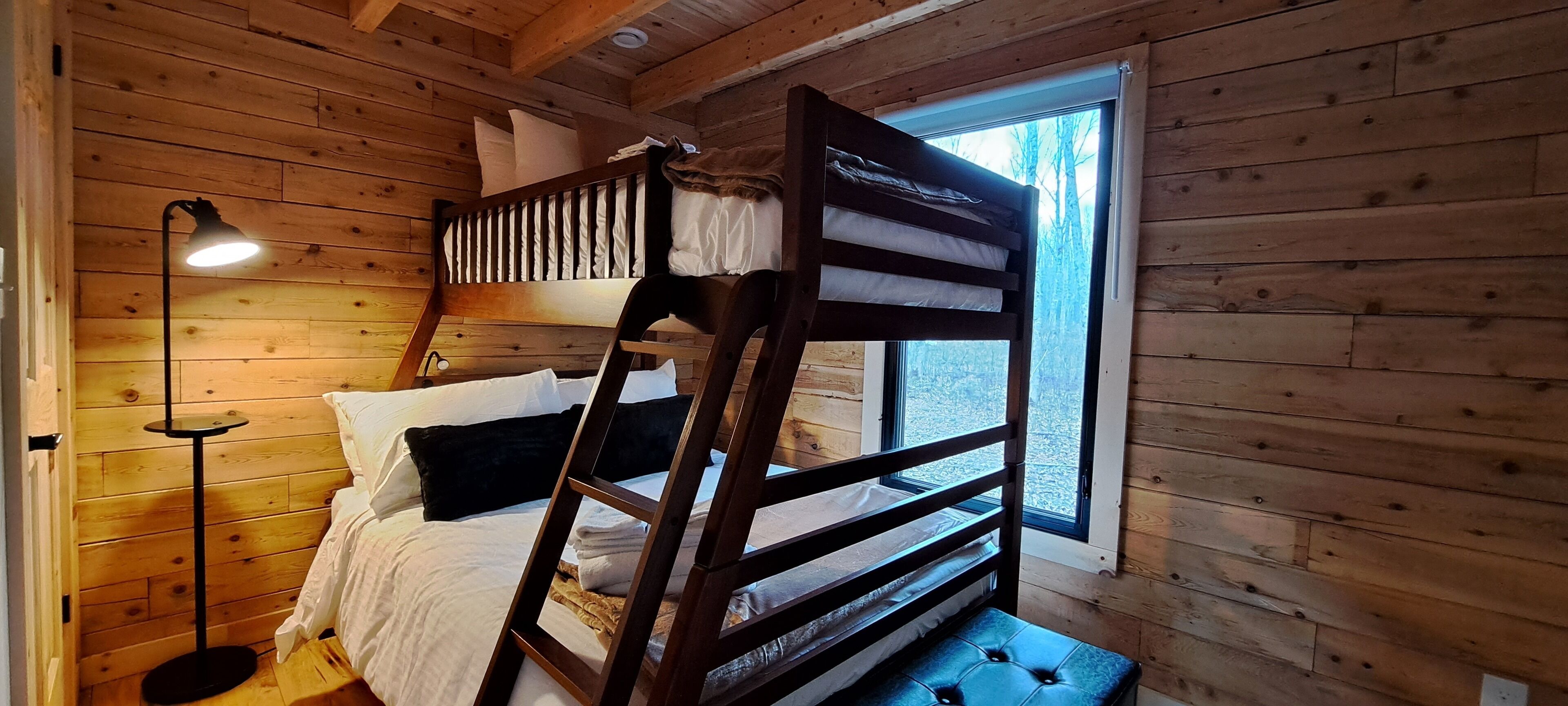 Een stapelbed met een ladder, een nachtkastje, een lamp en een raam met uitzicht op bomen.
