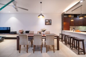 A modern dining area with a wooden table, four chairs, a ceiling fan, pendant lights, a television, and a kitchen counter with stools.