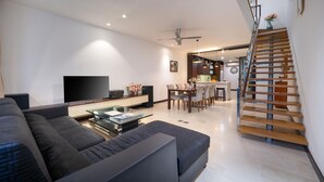 Una sala de estar moderna con televisor de pantalla plana, sofás oscuros, una mesa de centro de vidrio y una escalera que lleva a un nivel superior.