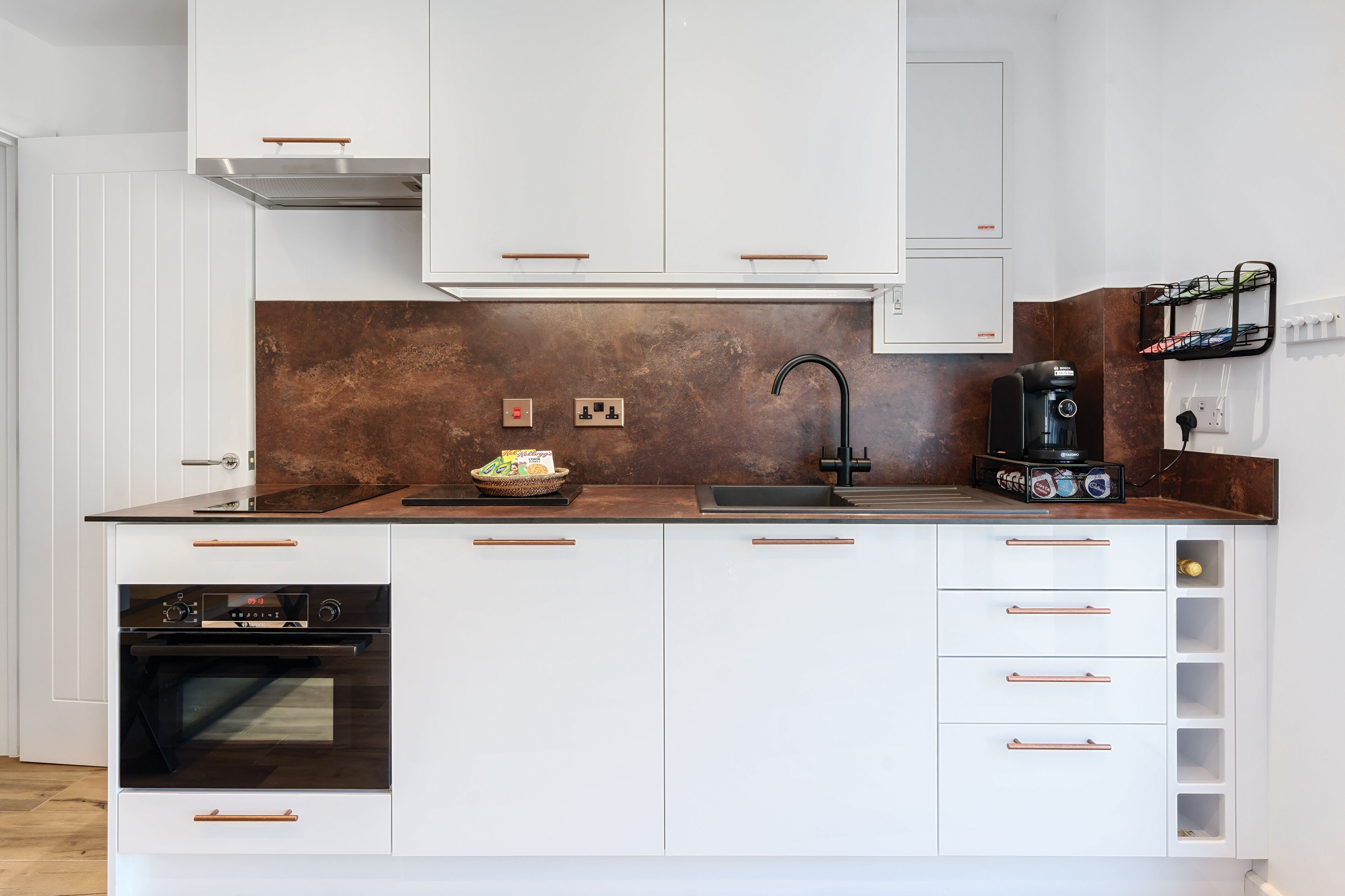 A modern kitchen with white cabinetry, a stainless steel range hood, a built-in oven, a coffee machine, and a backsplash with a brown tile pattern.