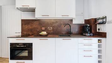 A modern kitchen with white cabinetry, a stainless steel range hood, a built-in oven, a coffee machine, and a backsplash with a brown tile pattern.