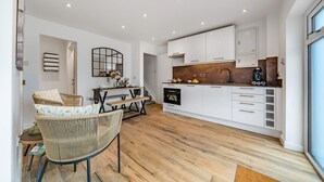 Een moderne keuken met witte kasten, een houten aanrechtblad en een ingebouwde oven. Er is een zithoek met rieten stoelen en een tafel, en een groot raam zorgt voor natuurlijk licht.