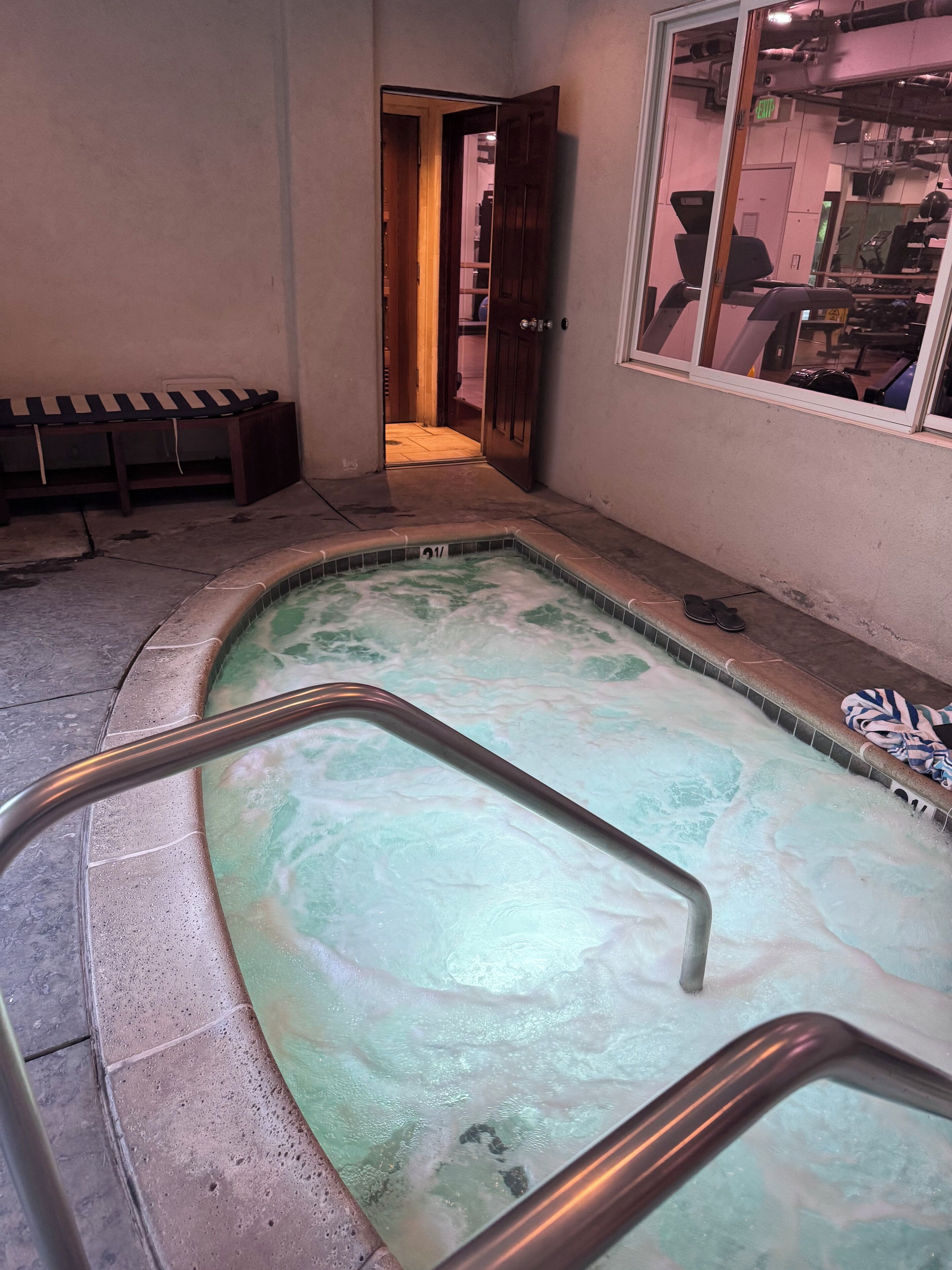 A small indoor pool with a metal ladder, a bench, and a door leading to another room.