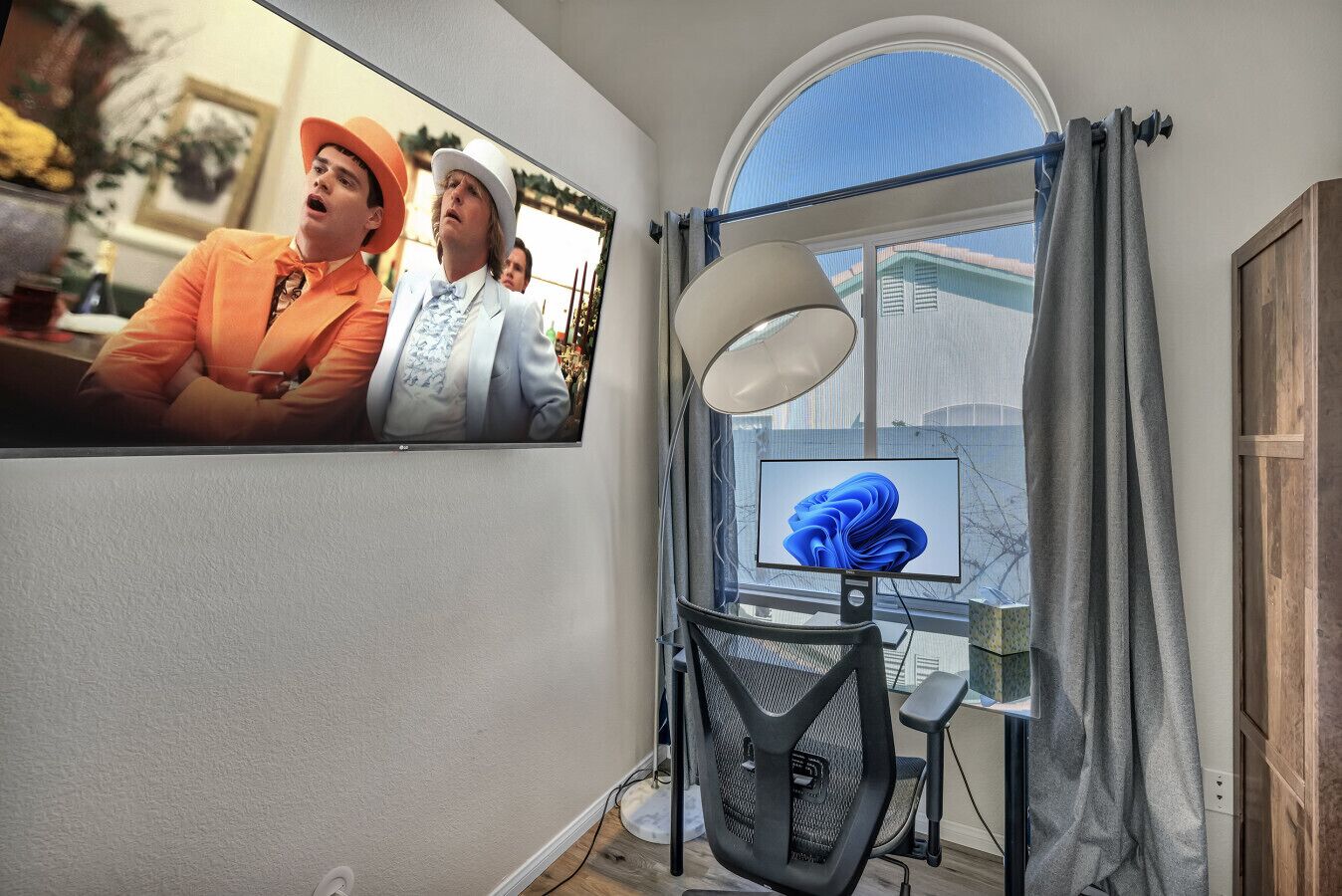 Habitación con una amplia televisión de pantalla plana montada en la pared, un escritorio con una silla, una ventana con cortinas y una lámpara.