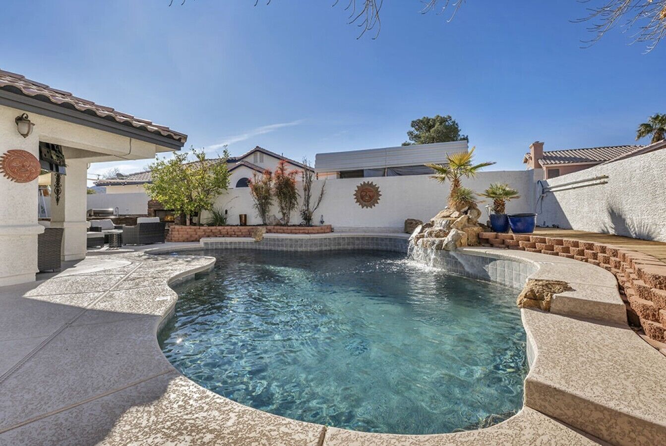 A clear swimming pool with a stone border, a small waterfall feature, and a patio area with outdoor furniture.