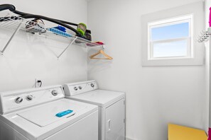 A laundry room with two white washers and dryers, a drying rack with clothes and a vacuum cleaner, a wooden hanger, and a window with a view.