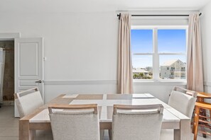A modern dining area with a rectangular table and six chairs, a wooden side table, and a view of buildings through a large window with beige curtains.