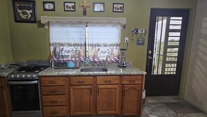 Una cocina con estufa, horno y alacenas de madera. Hay una ventana con cortina, y una puerta con ventanal.