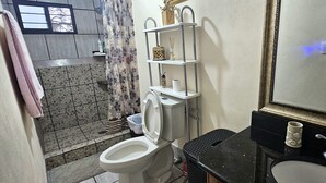 Un baño con inodoro, lavabo, espejo, repisa con artículos de aseo y zona de ducha.