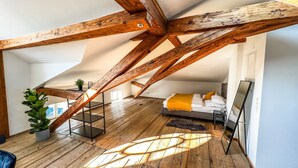 Une chambre à coucher aménagée en mezzanine avec des poutres apparentes, un lit agrémenté d’un plaid jaune et une grande fenêtre laissant entrer la lumière naturelle.