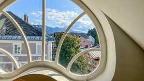 Une vue à travers une fenêtre à cadre incurvé, qui révèle une rue bordée d’immeubles et, au loin, une montagne.