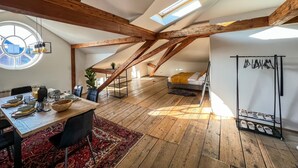 Une pièce de style « loft », avec des poutres en bois apparentes, une lucarne, un coin repas avec une table dressée pour quatre personnes et un lit recouvert d’une couverture jaune.