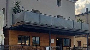 Un edificio de dos pisos con techo plano, un balcón con barandilla de vidrio y una entrada a nivel de calle con fachada de madera.
