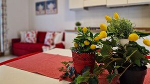 A living room with a red sofa, a coffee table with a red placemat, and two potted plants with yellow flowers. - Cozy Apartment With Swimming Pool (San Michele al Tagliamento)