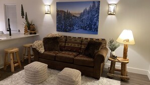 A cozy living room with a brown sofa, two ottomans, a fluffy rug, a wooden side table with a lamp, and a wall-mounted painting of a snowy forest. - The Comfy Bear Cottage! Beautiful 2 bed/1bath home minutes from skiing and lake. (Sandpoint)