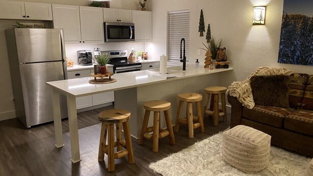 A modern kitchen with white cabinets, a stainless steel refrigerator, a microwave, and a coffee maker. There are wooden stools and a white rug in the living area. - The Comfy Bear Cottage! Beautiful 2 bed/1bath home minutes from skiing and lake. (Sandpoint)