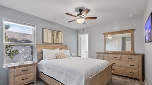A bedroom with a wooden bed, a ceiling fan, a large window with a view, and a wooden dresser.