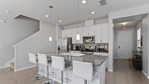 A modern kitchen with white bar stools, stainless steel appliances, and pendant lighting.