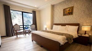Hotelkamer met een groot bed, een bureau en een stoel. Er is een raam met gordijnen, een schilderij aan de muur en uitzicht op een bergachtig landschap.