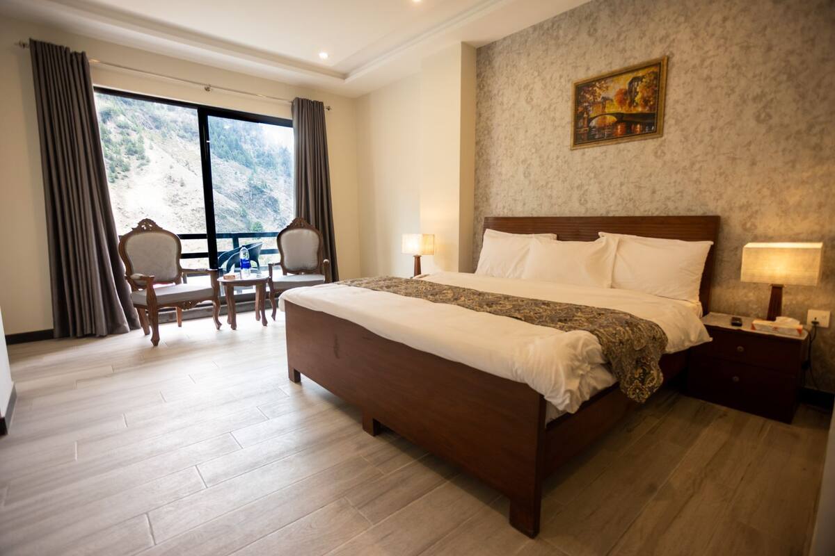 A hotel room with a large bed, a desk, and a chair. There is a window with curtains, a painting on the wall, and a view of a mountainous landscape.
