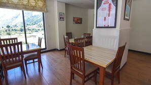 Een eethoek met houten tafels en stoelen, grote ramen met uitzicht op een bergachtig landschap, en een lijst met een poster aan de muur.