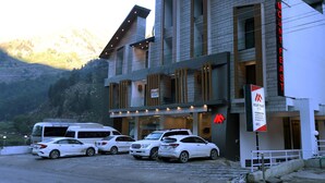 Een hotel met een modern design, met een opvallend bord voor 'Mount Feast Hotel'. Er zijn meerdere witte auto's geparkeerd voor het gebouw, dat tegen een bergachtige achtergrond ligt.