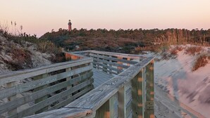 Ein hölzerner Steg führt zu einem Leuchtturm, umgeben von Dünen und spärlicher Vegetation.