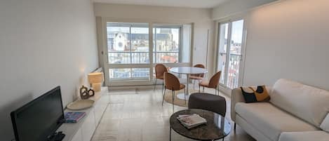 A modern living room with a flat-screen TV, a sofa, a coffee table, and a dining table with chairs. Large windows offer a view of buildings and a church steeple.