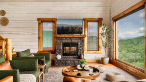 A cozy living room with a stone fireplace, green upholstered furniture, and a large window offering a scenic view.