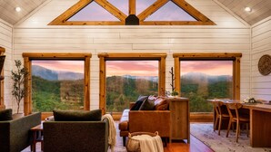 A cozy cabin interior with a wooden interior, large windows offering scenic mountain views, a dining area, and a seating area with a fireplace.