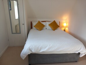 Een bed met witte lakens en twee gouden kussens, een bedlampje en een spiegel aan de muur.