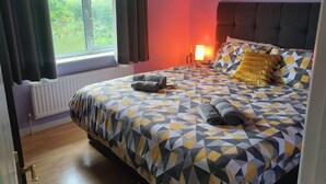A bedroom with a geometric patterned bedspread, a headboard, a bedside lamp, and a view of greenery through the window. - Private Entire House - 3 Bedrooms, Garden, Parking (Brighton)