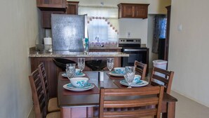 A dining area with a wooden table set for four, surrounded by chairs. A refrigerator and oven are visible in the background. - Captivating 2-bed House in Falmouth (Falmouth)