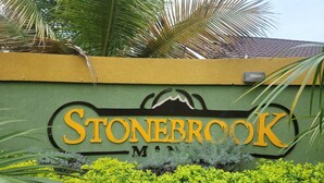 A wall with the name "Stonebrook" in large yellow letters, surrounded by green foliage and a palm tree. - Captivating 2-bed House in Falmouth (Falmouth)