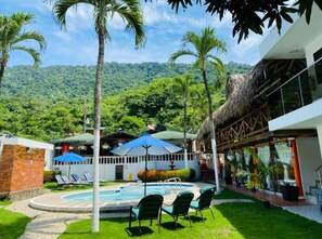 A pool area with lounge chairs, a thatched-roof structure, and a mountain backdrop. - hotel randuky tayrona  (Santa Marta)