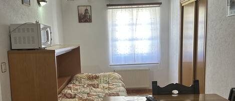 A compact room with a bed, a wooden table, a chair, a radiator, a window with curtains, and a small kitchenette area.