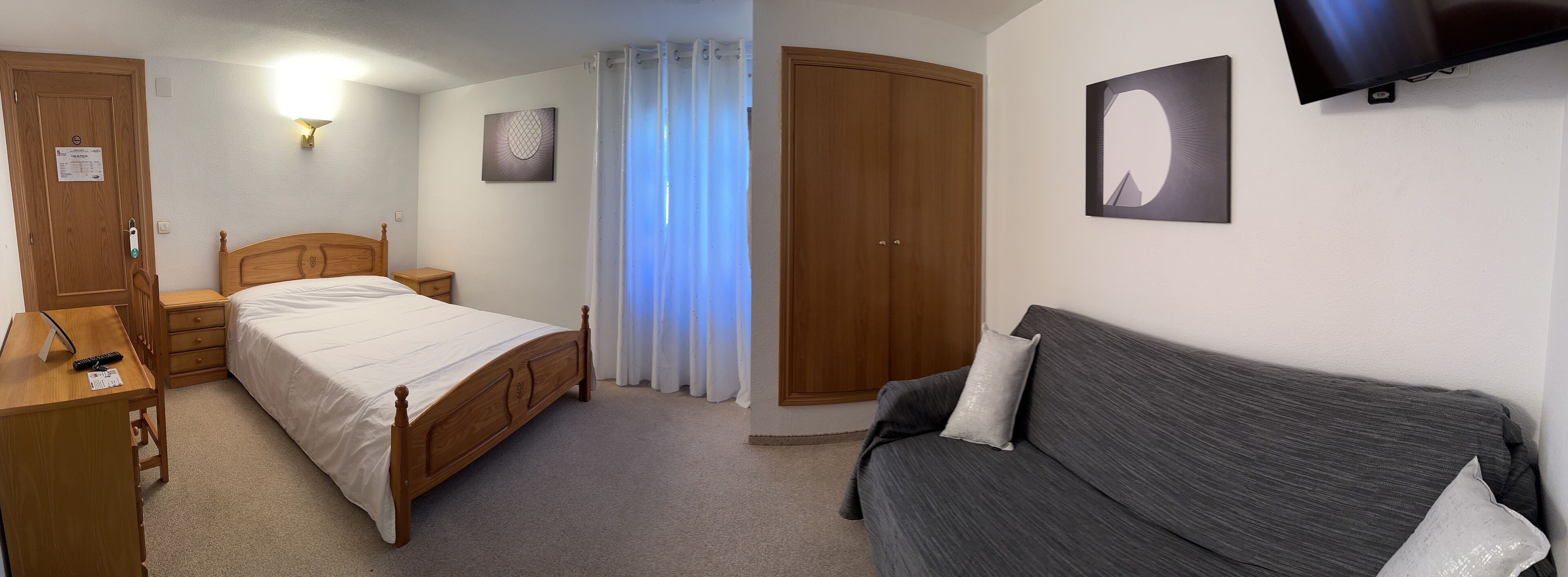 Un dormitorio con cama de madera, una mesita de noche, un escritorio con silla, un sofá gris, un armario y un televisor montado en la pared.