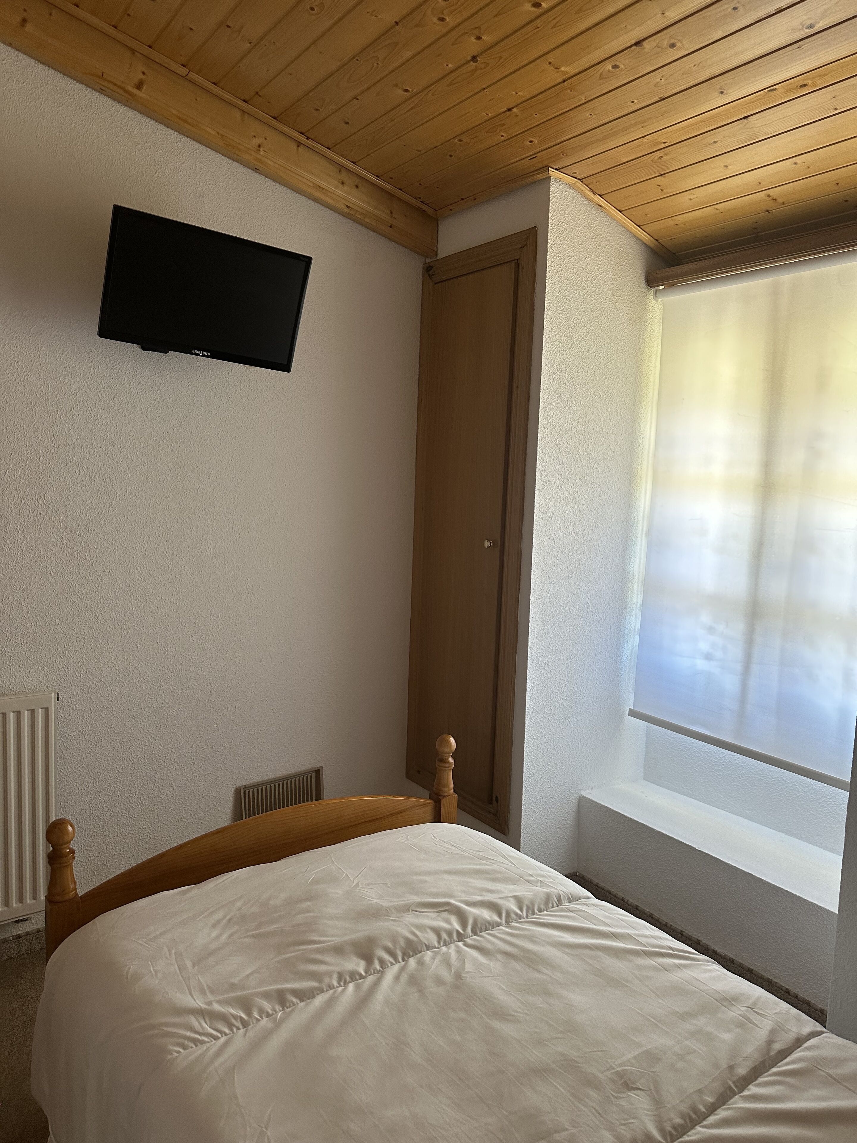 A bedroom with a wooden ceiling, a bed with a wooden headboard, a wall-mounted TV, a door, and a window with a view.