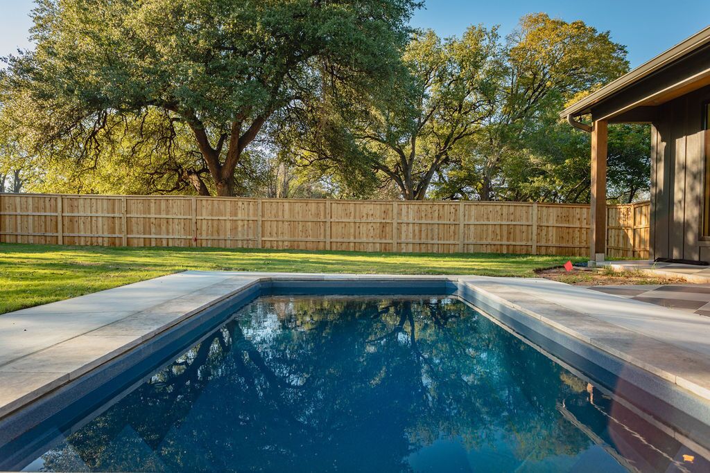 A rectangular swimming pool with a wooden deck surrounding it, a wooden fence, and trees in the background.