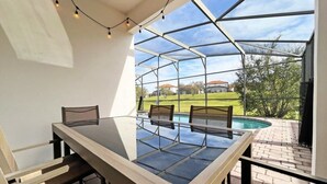 En täckt uteplats med ett glasbord och stolar, med utsikt över en swimmingpool och ett grönområde.