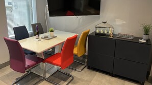 A modern office space with a large flat-screen TV mounted on the wall, a white table with red and yellow chairs, a black filing cabinet, and a small potted plant. - Modern house with a Tub!!! (Bangor)