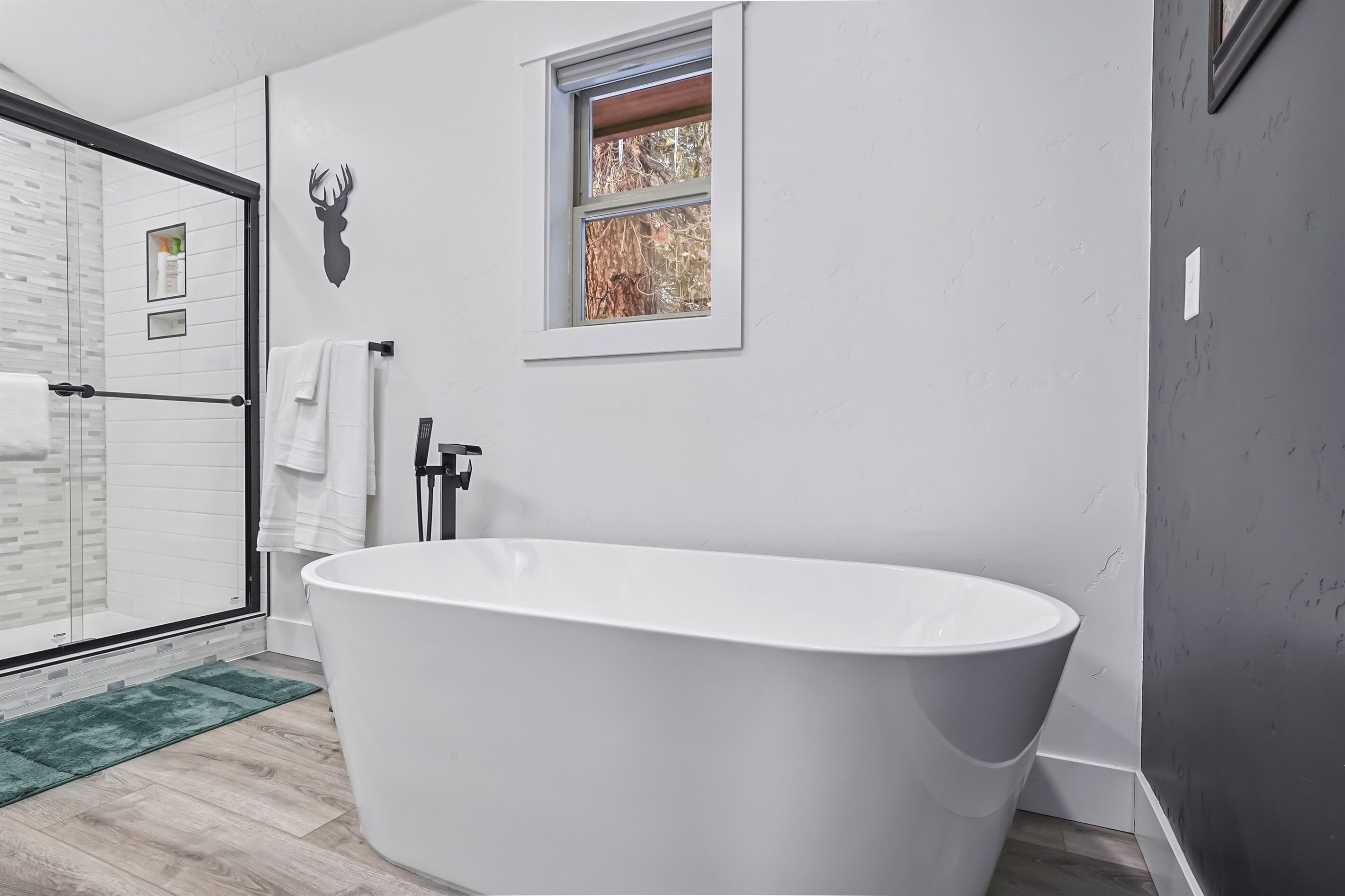 A modern bathroom with a freestanding bathtub, a glass shower enclosure, and a window with a view of trees.
